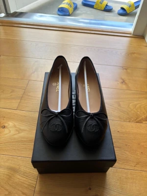 Svarta ballerinaskor från Chanel - Säljer ett par klassiska svarta ballerinaskor från Chanel i läder. Skorna har rund tå, dekorativ rosett framtill och den ikoniska Chanel-loggan broderad på tån. Insidan är beige och de kommer i originalkartong. Perfekta för en stilren och elegant look.