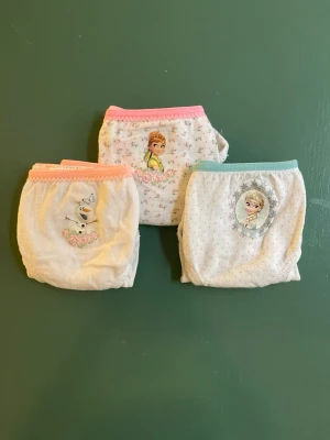 Frost trosor med motiv, 3-pack - Säljer ett 3-pack trosor med motiv från Disney Frost. Trosorna är vita med små blommor och har olika karaktärer framtill, bland annat Elsa, Anna och Olaf. Resår i midjan i rosa eller turkos. Perfekta för dig som gillar Frost och vill ha söta underkläder.