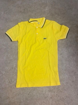 Gul pikétröja från Lacoste - Snygg gul pikétröja från Lacoste med klassisk krage och två knappar framtill. Tröjan har korta ärmar med vita och svarta detaljer på krage och ärmslut samt den ikoniska gröna krokodilloggan på bröstet. Perfekt för en fräsch och sportig look.