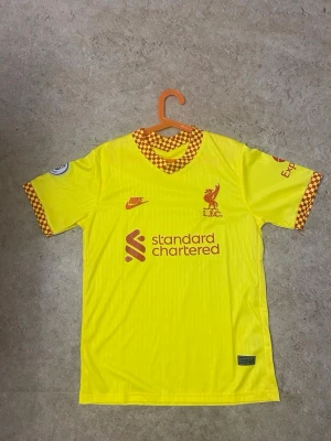 Liverpool FC gul matchtröja Nike - Gul Liverpool FC matchtröja från Nike med röda detaljer och rutigt mönster på krage och ärmslut. Tröjan har klubbmärke, Nike-logga och sponsortryck framtill. Tillverkad i lätt och ventilerande material, perfekt för fotboll eller träning.