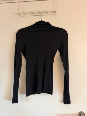 Svart ribbad polotopp - En svart, ribbad polotopp med lång ärm. Toppen har en tight passform och är tillverkad i ett mjukt stretchigt material som sitter snyggt på kroppen. Perfekt att styla med jeans eller kjol för en clean look. Aldrig använd. Köpt på vinted