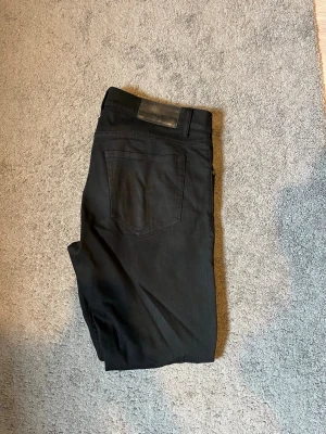 Svarta jeans från Tiger of sweden - Säljer ett par svarta slim fit jeans från Tiger of sweden i modellen Evolve med klassisk femficksdesign och diskreta detaljer. Jeansen har en svart läderpatch bak i midjan och metallknappar framtill. Materialet är stretchigt för extra komfort och passform.