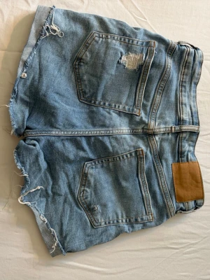 Ljusblå slitna jeansshorts - Säljer ett par ljusblå jeansshorts med råa, fransiga kanter och slitningar både fram och bak. Klassisk femficksmodell med dragkedja och knapp. Perfekta för en avslappnad och trendig sommarstil.