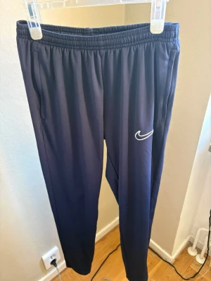 Mörkblå Nike träningsbyxor  - Säljer ett par mörkblå träningsbyxor från Nike med vit broderad logga på benet. Byxorna har resår i midjan och smal passform med fickor på sidorna. Perfekta för chill eller träning, tillverkade i svalt och bekvämt material.