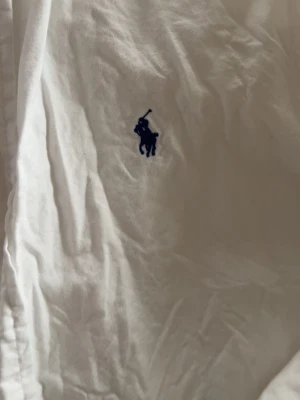 Vit skjorta från Ralph Lauren - Klassisk vit skjorta från Ralph Lauren med den ikoniska blå broderade loggan på bröstet. Skjortan har knappar framtill och lång ärm. Tillverkad i mjuk bomull och passar perfekt till en clean och stilren look.