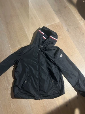 Svart vindjacka - Snygg svart vindjacka med huva och dragkedja framtill. Jackan har vita och röda detaljer på huvan samt två fickor med tryckknappar. Moncler-logga på ärmen. Tillverkad i lätt, glansigt material som står emot vind och regn. Har bara använt 2 gånger och säljer på grund av att den är för stor.