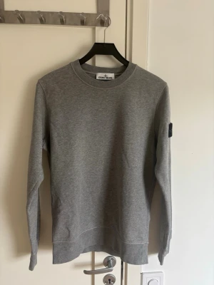 Grå sweatshirt från Stone Island - Snygg grå sweatshirt från Stone Island med klassisk rund hals och ribbade muddar. Tröjan har den ikoniska Stone Island-loggan på vänster ärm och är tillverkad i mjukt bomullsmaterial. Perfekt för en avslappnad och stilren look.