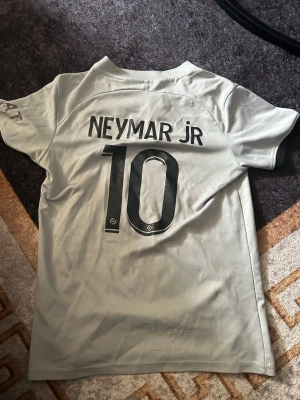 PSG Neymar Jr matchtröja grå - Snygg grå PSG matchtröja med Neymar Jr och nummer 10 på ryggen. Framtill finns PSG-logga, Jordan-logga och Qatar Airways tryck. Tröjan har korta ärmar och svart rand längs framsidan. Perfekt för fotbollsfans och streetwear.