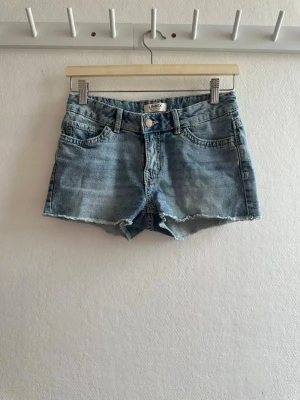 Blå jeansshorts från Lindex - Snygga blå jeansshorts från Lindex med rå kant nertill och klassisk femficksdesign. Shortsen har knapp och dragkedja framtill samt dekorativa bakfickor med lock och knapp. Perfekta för varma dagar.