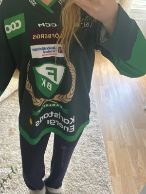 Färjestad BK hockeytröja grön CCM - Säljer en grön hockeytröja från Färjestad BK med stora loggor och sponsormärken framtill. Tröjan har långa ärmar, V-ringning och detaljer i vitt och guld. Perfekt för dig som vill stötta laget på match eller till vardags. ALDRIG ANDVÄND. Köpte för 500 kr.