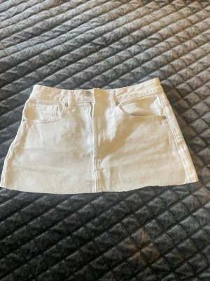 Vit kort jeanskjol med fickor - Säljer en vit kort jeanskjol med klassisk femficksdesign och rå kant nertill. Kjolen har bälteshällor och stängs med knapp och dragkedja framtill. Perfekt till sommaren och enkel att matcha med olika toppar.
