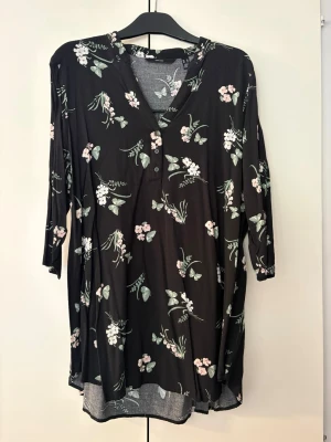 Svart blommig blus Vero Moda trekvartsärm - Svart blus från Vero Moda med blommor och fjärilar i grönt och rosa. V-ringning med knappslå framtill och trekvartsärmar. Lätt och mjukt material som ger en avslappnad passform. Perfekt för dig som gillar romantiska mönster och sköna plagg. Storlek XL.
