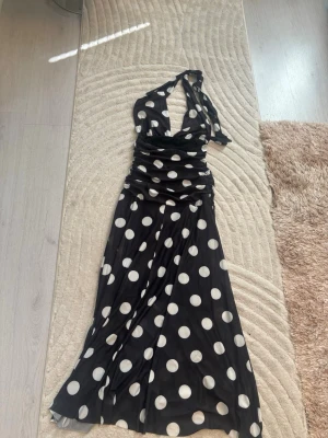 Svart halterneckklänning med prickar - Snygg lång svart klänning med stora vita prickar och halterneck. Klänningen har djup urringning och markerad midja med rynkade detaljer. Tyget är mjukt och följsamt, perfekt för en trendig look.