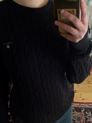 GANT cable knit sweater svart – klassisk stickad tröja - Klassisk svart/mörk navy kabelstickad tröja från GANT med broderad logga på bröstet.  Rund hals, ribbade muddar och fin preppy/old money-känsla. Superfin till jeans, skjorta, kostymbyxor eller kjol.  Fint begagnat skick med normala små bruksspår/ludd, se bilder.  Storlek: M Material: 100% bomull Tyg: Stickat Skötselråd: Torktumlas ej