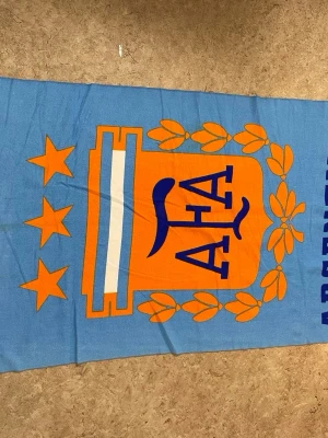 Argentina strandhandduk blå och orange - Stor strandhandduk med Argentinas fotbollsförbunds logga i blått och orange. Handduken har tre stjärnor, lagerkrans och texten 'ARGENTINA' längs kanten. Perfekt för stranden eller poolen, tillverkad i mjukt material.