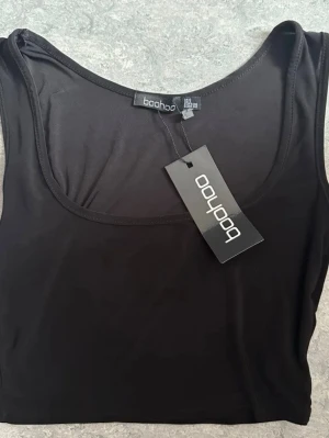 Svart croppad topp Boohoo - Snygg svart croppad linnetopp från Boohoo med rundad halsringning. Tillverkad i ett stretchigt syntetmaterial som sitter tight mot kroppen. Perfekt att styla till jeans eller kjol för en clean look.