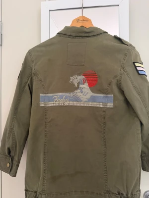Zadig Voltaire jacka ( KAN GÅ NER I PRIS) - (KAN GÅ NER I PRIS SÅ BUDA KOMMER NOG GODKÄNNA).                    Snygg olivgrön Zadig Voltaire jacka  med broderad våg och röd sol på ryggen samt coola patchar på ärmarna. Jackan har flera fickor med knappar och dragkedjor framtill, axelklaffar och justerbar midja. Perfekt för dig som vill sticka ut med detaljer. Strl xs