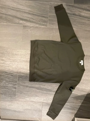 Olivgrön sweatshirt från Stone Island - Säljer en stilren olivgrön sweatshirt från Stone Island med klassisk rund halsringning och ribbade muddar. Tröjan har den ikoniska Stone Island-loggan på vänster ärm. Perfekt för dig som gillar streetwear och enkel design. Är size m men passar s