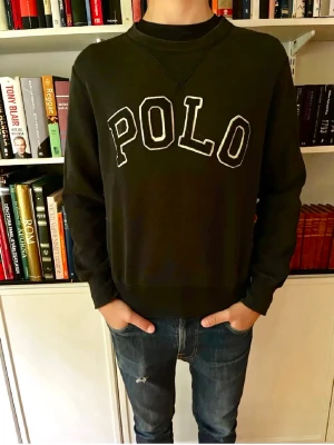 Svart sweatshirt från Polo Ralph Lauren - Cool svart sweatshirt från Polo Ralph Lauren med stor vit POLO-text på bröstet. Tröjan har rund halsringning, ribbade muddar och avslappnad passform. Perfekt till jeans för en casual look.