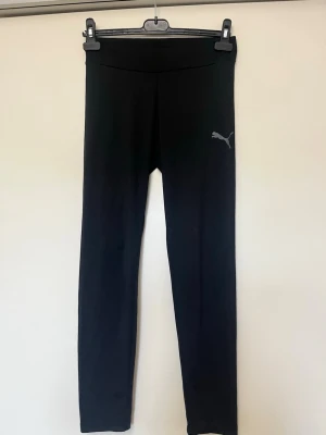 Svarta sport leggings från Puma - Snygga svarta leggings från Puma med klassisk logga på låret. De har hög midja och är tillverkade i ett stretchigt material som sitter skönt på kroppen. Perfekta för träning eller chill. Tight passform och stilren design.