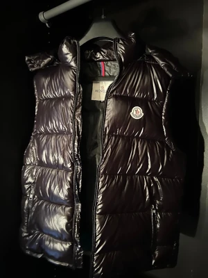 Svart dunväst från Moncler - Snygg svart dunväst från Moncler med glansig finish och klassisk logotyp på bröstet. NFC tillgängligt på loggan. Västen har dragkedja framtill, hög krage och två fickor med dragkedja. Perfekt för lager-på-lager och streetstyle. Materialet är dunfyllt och yttermaterialet är blankt.