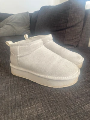 Vita fluffiga tofflor med platåsula - Mysiga vita tofflor med hög platåsula och mjuk, fluffig insida. Utsidan är i mockaliknande material och tofflorna har dragflik bak för enkel påtagning. Perfekta för att hålla fötterna varma hemma under kyliga dagar. Finns en färgfläck på i turkos därav priset.
