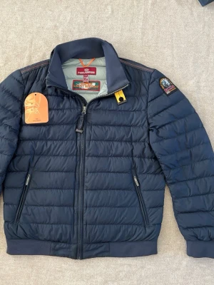 Mörkblå dunjacka från Parajumpers - Snygg mörkblå dunjacka från Parajumpers med quiltad design, hög krage och dragkedja framtill. Jackan har två sidofickor med dragkedja och en ikonisk patch på ärmen. Perfekt för kalla vinterdagar och riktigt stilren look.