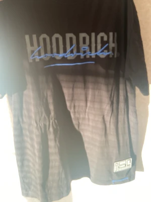 Svart t-shirt från Hoodrich - Snygg svart t-shirt från Hoodrich med stort tryck i vitt och blått framtill och baktill. T-shirten har rund hals, korta ärmar och en relaxed passform. Tillverkad i mjuk bomull som känns skön mot huden. Perfekt för en streetwear-look.