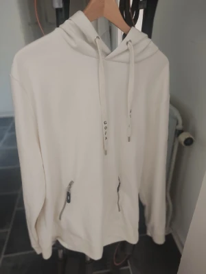 Vit hoodie från GOJA med dragkedjor - Snygg vit hoodie från GOJA med huva och dragsnören. Två praktiska fickor med dragkedja framtill. Tillverkad i mjukt material som känns skönt mot huden. Perfekt för en avslappnad och stilren look. Nypris 1500:-