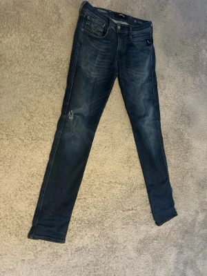 Mörkblåa andbass jeans från Replay - Säljer ett par blå slim fit jeans från Replay, modell Hyperflex Anbass. Jeansen har klassisk femficksdesign, stretchigt material och snygga slitningar på benen. Perfekta för dig som gillar en smal passform och vill ha bekväma jeans med cool tvätt.