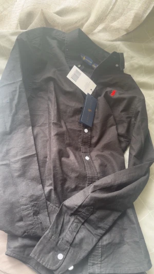 Svart skjorta från Ralph Lauren - 10/10 nytt skick Klassisk svart skjorta från Ralph Lauren med röd broderad logga på bröstet. Skjortan har långa ärmar, vita knappar och en tidlös design som passar till mycket. Tillverkad i bomull för en skön känsla och snygg passform.