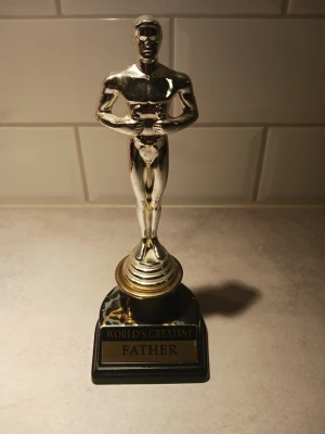 World's Greatest Father statyett - Rolig statyett i guld- och svartfärgad plast med texten 'WORLD'S GREATEST FATHER' på en skylt. Perfekt som present till pappa eller som dekoration. Statyetten har en klassisk pokalform och står på en svart fot.