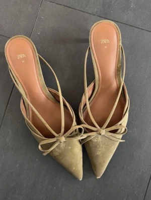 Olivgröna pumps med rosett från ZARA - Säljer ett par eleganta olivgröna pumps från ZARA med spetsig tå och tunna remmar. Skorna har en dekorativ rosett framtill och är tillverkade i ett material som liknar mocka. Perfekta för dig som vill ha en stilren och trendig look.
