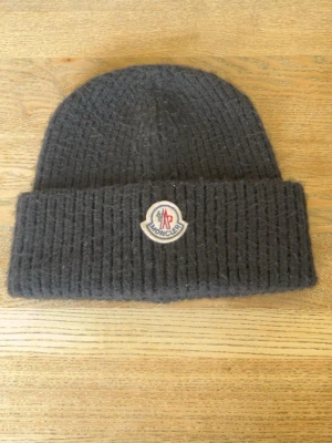 Mörkgrå stickad Moncler mössa - Säljer en mörkgrå stickad mössa från Moncler med uppvikt kant och klassisk Moncler-logga framtill. Mössan är ribbstickad och har en mjuk, ullig känsla som håller dig varm under kalla dagar. Perfekt accessoar för dig som vill ha både stil och komfort.