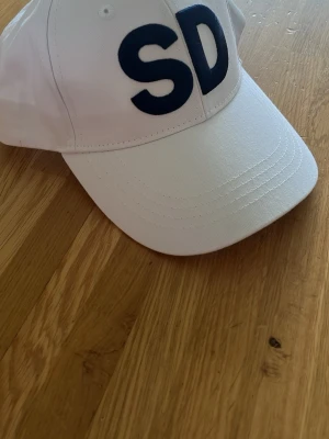 SD keps - Säljer en stilren Sverige Demokraterna keps med stora, mörkblå bokstäver 'SD' broderade framtill. Klassisk böjd skärm och justerbar passform bak. Perfekt accessoar för en sportig och avslappnad look. Materialet är bekvämt.
