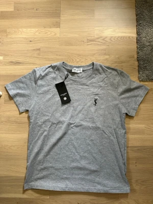 Saint Laurent T shirt - Grey - Tja! Säljer en riktigt stilig ysl t shirt med tags. Skick 10/10 allt på bilden följer med! Den är perfekt till sommaren. Storlek L men passar som en M. Skriv hur lång du är så kan jag räkna ut om den passar. Pris 399kr går att diskuteras vid en snabb affär. Postar med PostNord! MVH Etienne 
