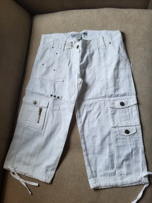 Vita cargobyxor med snörning Wet Seal Storlek 7US M - Säljer ett par vita cargobyxor med flera fickor, dragkedjedetalj och knappar. Byxorna har snörning vid bensluten och avslappnad passform. Perfekta för en chill och street-inspirerad stil. Materialet känns som bomull och byxorna har klassiska cargodetaljer.