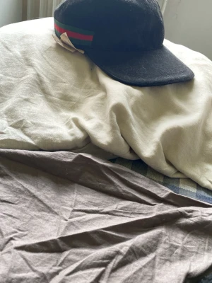Gucci - Säljer en svart keps från Gucci med klassisk röd och grön rand på sidan. Kepsen har böjd skärm och är tillverkad i ett mjukt material som känns lyxigt. Insidan har märkning och etikett från Gucci. Perfekt för dig som gillar exklusiva accessoarer.