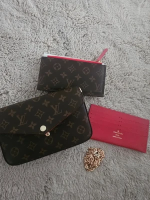  monogram plånbok set - Säljer ett set plånböcker i klassiskt brunt monogrammönster och detaljer i rött. Setet innehåller en kuvertformad plånbok med guldfärgad knapp, en zip-pouch med röd dragkedja och ett rött kortfodral i läder. Perfekt för dig som gillar lyxiga accessoarer.