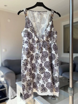 Vit blommig sommarklänning - Söt vit klänning med grått blommönster och djup v-ringning. Ärmlös modell med breda axelband och lätt a-linjeformad passform. Perfekt för varma dagar och enkel att styla med sandaler eller sneakers.