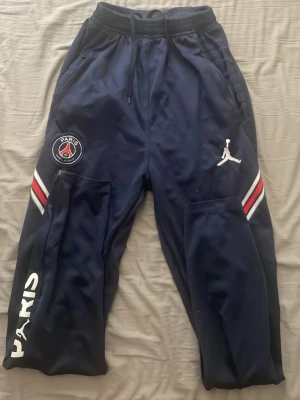 PSG x Jordan marinblå träningsbyxor - Snygga marinblå träningsbyxor från PSG x Jordan med vita och röda detaljer på benen. Byxorna har resår i midjan med dragsko, sidofickor och dragkedjor vid bensluten. PSG-logga på framsidan samt PARIS-tryck på benet. Jättesköna och går att använda till vardags också. Minns tyvärr ej vart jag köpte de från eller vad de kostade nya. Jag har för mig att dom är storlek S. Passar även mig som har storlek S. 
