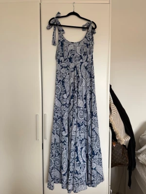Blåvit långklänning med knytband - Långklänning i blått och vitt med paisleyinspirerat mönster. Klänningen har knytband på axlarna och en luftig, svepande passform. Perfekt för varma dagar och somriga tillfällen.