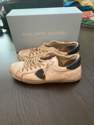 Philippe Model vita sneakers - Snygga vita sneakers från Philippe Model med svart detalj på sidan och svart hälparti. Skorna har klassisk låg profil, vita snören och är tillverkade i läder. Perfekta för dig som gillar stilrena och trendiga sneakers.