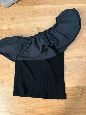Svart one shoulder topp med volang - Trendig svart one shoulder topp med bred volang runt axlarna. Toppen är ribbad och har en croppad passform som passar perfekt till höga jeans eller kjol. Perfekt för dig som vill ha en stilren men ändå iögonfallande look. Har köpt på vinted så vet tyvärr inte vad för märke då det inte finns någon tydlig lapp.