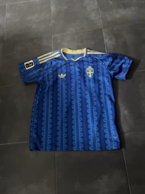 Sverige landslagströja - Säljer en blå fotbollströja från Adidas med Sveriges landslagsemblem på bröstet. Tröjan har gula detaljer, tre gula ränder på axlarna och ett unikt mönster över hela tyget. Perfekt för dig som vill stötta Sverige på match eller träning.