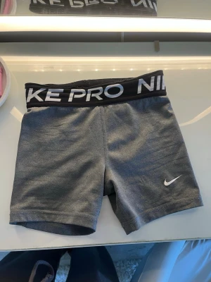 Grå Nike Pro träningsshorts - Säljer ett par grå Nike Pro träningsshorts med svart resår i midjan och vit Nike-logga på benet. Shortsen är tillverkade i ett stretchigt och mjukt material som passar perfekt för träning och sportaktiviteter. Snygg och sportig design med tight passform.storlek M BARN
