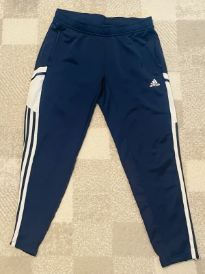 Mörkblå Adidas träningsbyxor - Snygga mörkblå träningsbyxor från Adidas med vita detaljer och klassiska ränder längs sidorna. Byxorna har smal passform, resår i midjan och dragkedjefickor. Tillverkade i ett mjukt och stretchigt material som passar perfekt till träning eller chill.