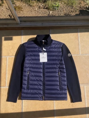 Mörkblå cardiganjacka från Moncler - Snygg mörkblå cardiganjacka från Moncler med quiltad framsida och stickade ärmar samt rygg. Jackan har två dragkedjefickor fram, hög krage och Moncler-logga på ärmen. Perfekt för lager-på-lager och stilren look.