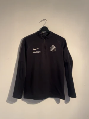 Svart AIK träningströja  - Svart långärmad träningströja från Nike med Dri-Fit material. Tröjan har AIK-logga på bröstet, vit Nike-swoosh och 'stadium'-tryck. Praktisk half zip-dragkedja vid halsen och sportig passform. Perfekt för fotbollsträning eller annan sport. Storlek XL (barnstorlek) passar xs i vanlig