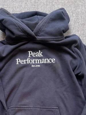 Mörkblå hoodie Peak Performance - Snygg mörkblå hoodie från Peak Performance med stor logga och texten 'Est. 1986' på bröstet. Klassisk huva och känguruficka framtill. Tillverkad i mjukt bomullsmaterial som känns skönt mot huden. Perfekt för en avslappnad och sportig stil.